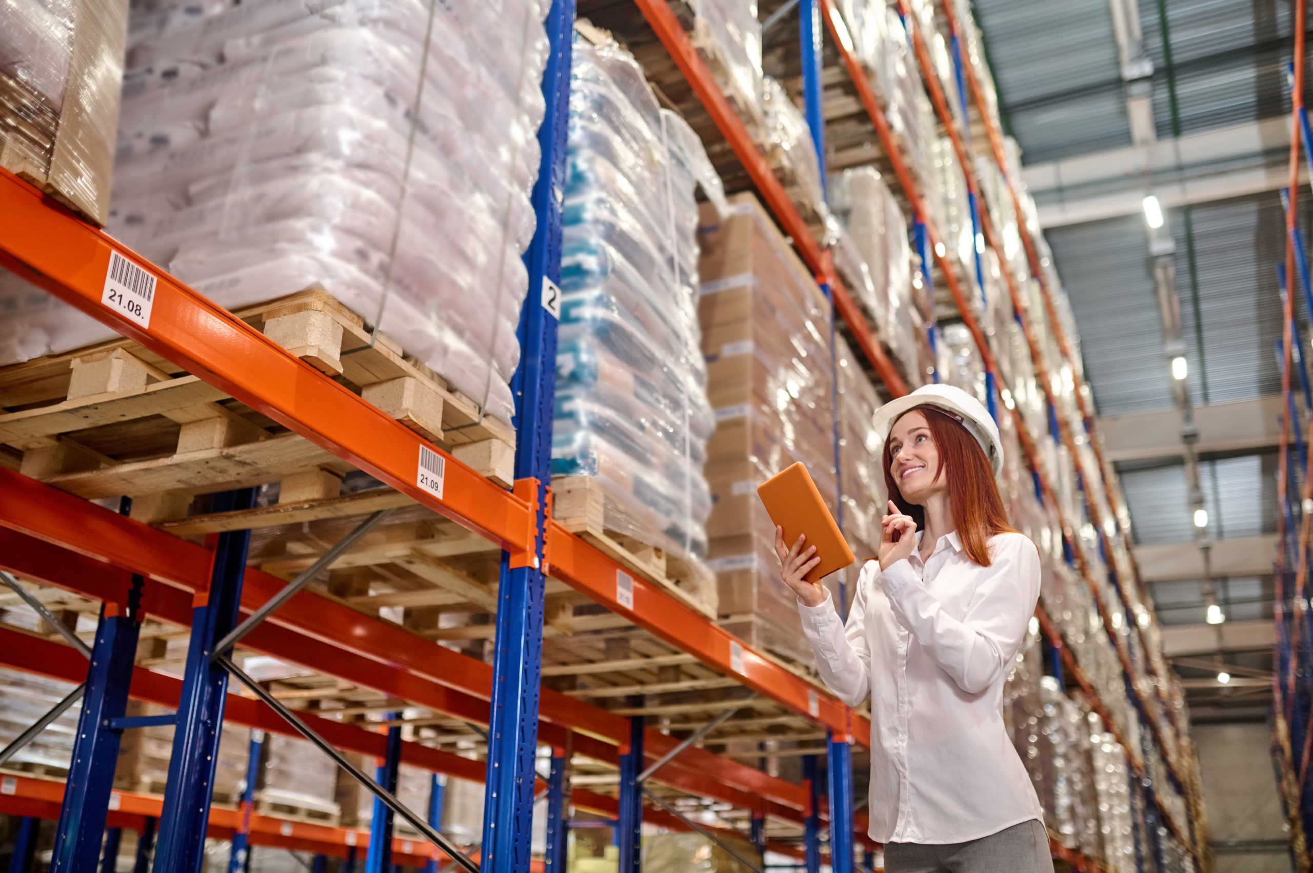 Warehouse, audit. Concentrated young woman with long red hair in safety helmet with tablet working checking near warehouse shelves with cargo